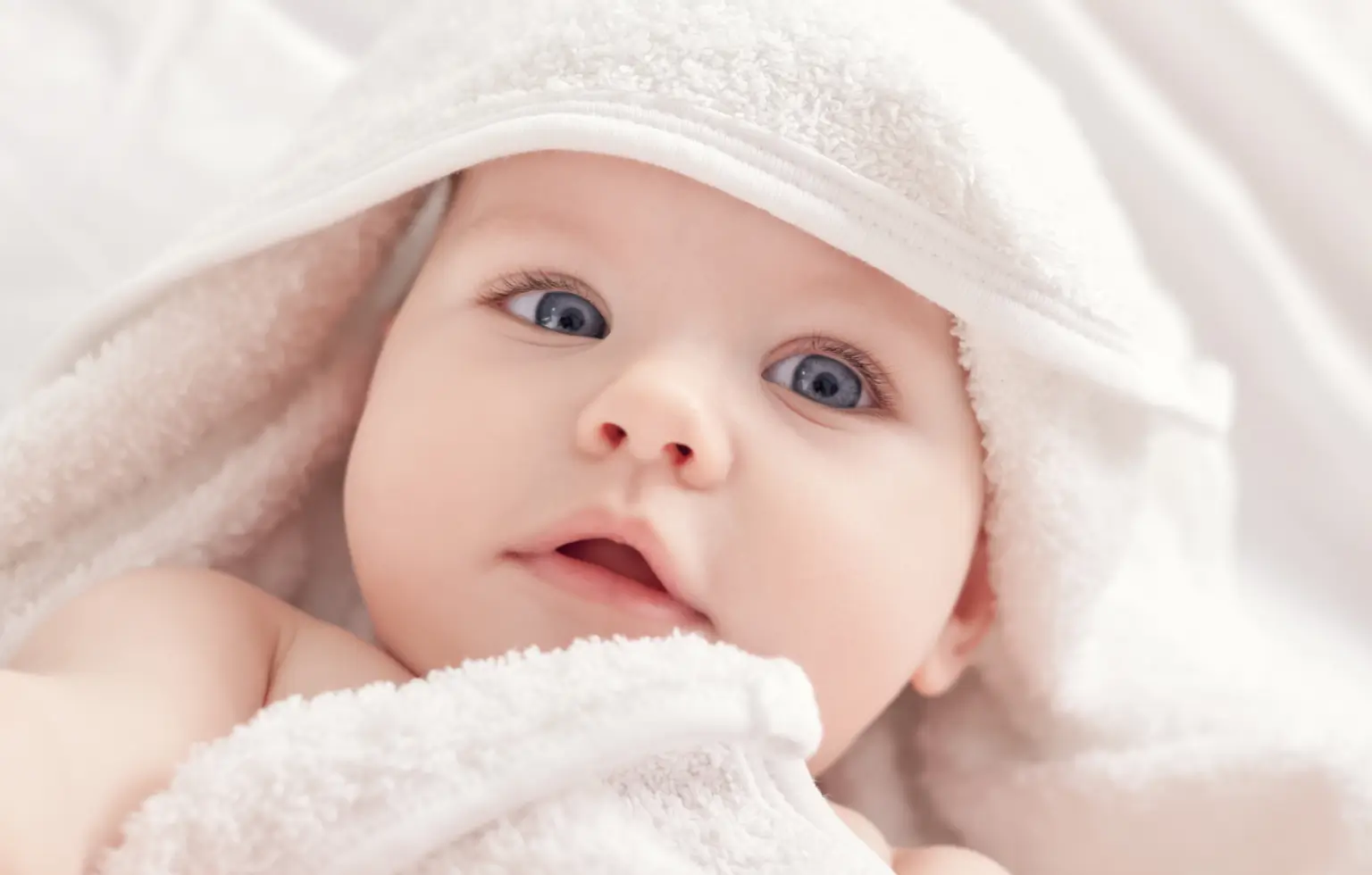 Bebê de olhos azuis enrolado em uma toalha branca felpuda.