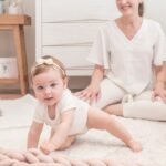 Bebê engatinhando em tapete com mãe ao fundo